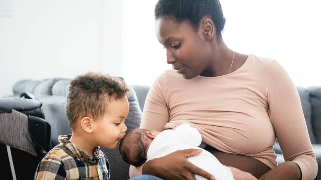 Black African American woman breastfeeding baby; toddler kissing baby's head. I