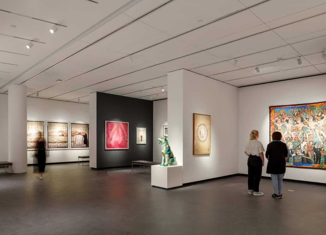 A modern museum gallery is photographed at a wide angle. It features several inset bays in which art of various sizes and mediums is hung. Visitors walk through the galleries and observe the works.