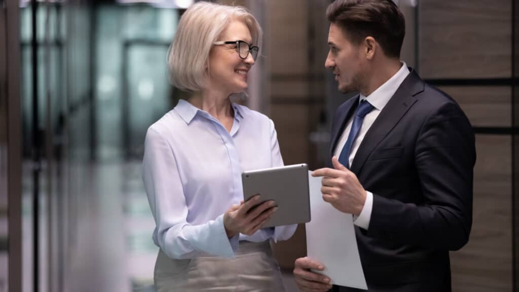 Smiling mature blonde female boss in glasses showing project details to younger male colleagues, standing together in office hall. Happy different ages workers discussing working issues in corridor.