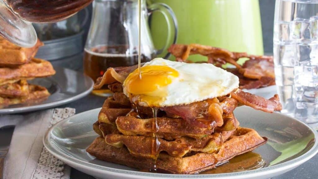 Cheddar-Waffles-stacked-with-bacon-and-egg-and-maple-syrup-poured-on-top-all-on-a-grey-plate.