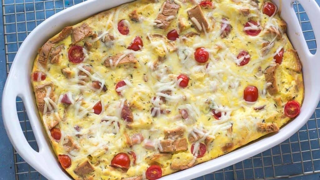 overhead-shot-of-ham-and-cheese-strata-in-a-white-casserole-dish-on-a-rack-against-a-blue-background.