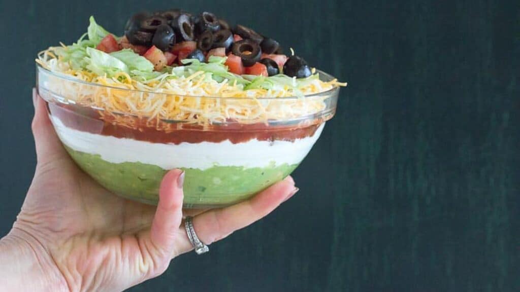 hand-holding-glass-bowl-with-7-layer-dip-against-green-crackled-background.