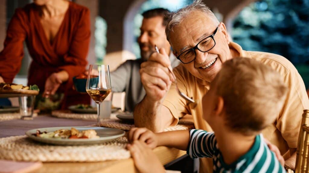 Happy grandfather having fun while feeding his grandson at dining table.