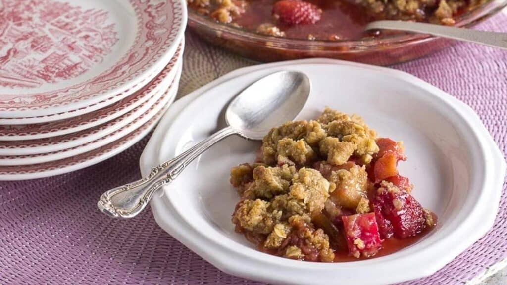 strawberry-rhubarb-crisp-on-a-white-plate-with-silver-spoon-stack-of-pink-plates-and-whole-crisp-in-background.