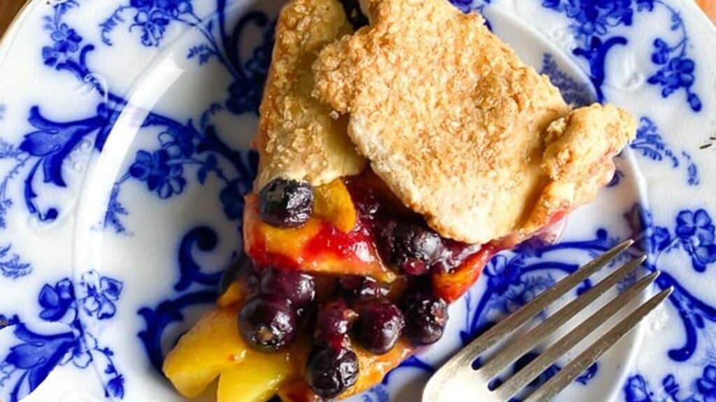 vertical-image-of-wedge-of-peach-blueberry-crostata-on-blue-and-white-plate-held-by-womans-hand.